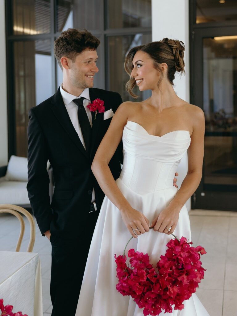 Newlywed couple smiling at each other while holding a heart-shaped pink floral bouquet during an elegant Naples wedding celebration coordinated by a Naples wedding planner and photographed by Priscila del Cristo.