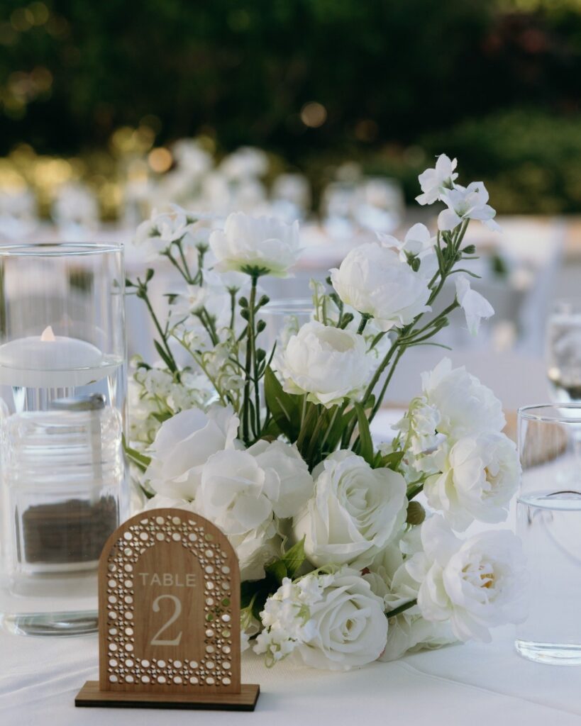 A refined white floral centerpiece featuring roses, ranunculus, and delicate greenery creates a timeless reception aesthetic planned by experienced wedding planners in Naples Florida. Thoughtful tabletop styling enhances the elegant guest experience at this beautifully curated Naples wedding. Photography by Priscila del Cristo.