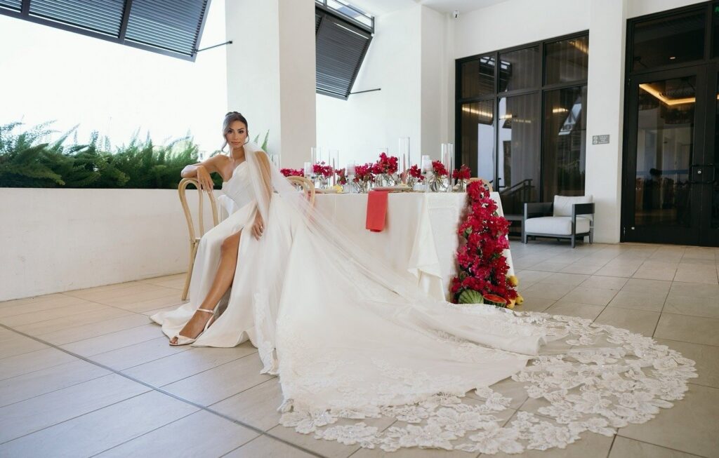 Bride seated beside an elegantly styled reception table featuring vibrant red floral arrangements and modern décor, with her long lace train flowing across the floor in a sophisticated Naples wedding setting. Styled editorial image showcasing the refined work of wedding planners in Naples, photographed by Priscila del Cristo, highlighting luxury design inspiration from top wedding planners Naples FL.