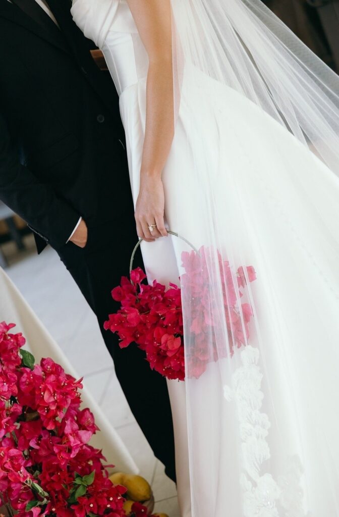 Close-up detail of a bride in a modern strapless wedding gown holding a vibrant pink floral bouquet beneath a flowing veil beside the groom’s black suit at a stylish Naples Florida wedding designed by expert wedding planners in Naples and photographed by Priscila del Cristo.