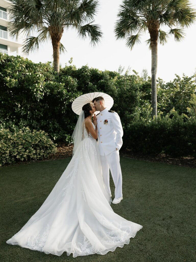 Bride and groom share a romantic kiss under a white parasol surrounded by tropical greenery and palm trees during an elegant outdoor Naples wedding planned by a Naples Florida wedding planner and captured by photographer Priscila del Cristo.