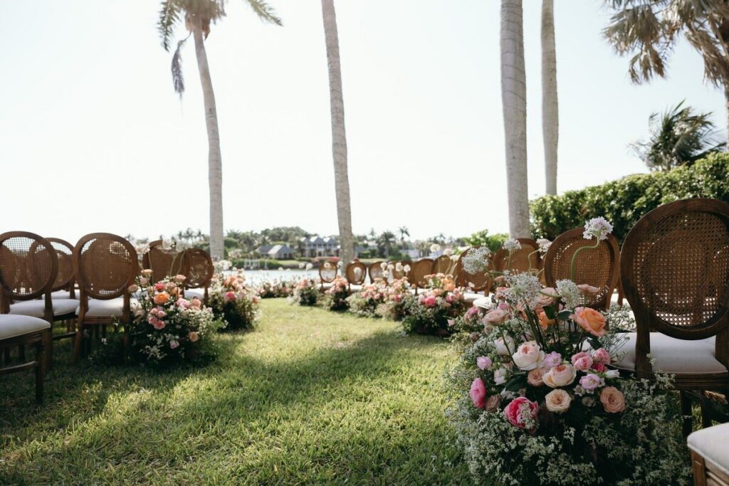 This beautifully styled outdoor ceremony showcases a waterfront garden aisle lined with lush floral arrangements in soft blush, peach, and ivory tones. Cane-back ceremony chairs frame the aisle, creating an intimate and welcoming atmosphere beneath towering palm trees overlooking the Naples shoreline. Thoughtfully curated layouts like this demonstrate how experienced wedding planners in Naples Florida transform natural coastal venues into refined ceremony spaces that feel effortless and luxurious. From ceremony flow and guest seating placement to floral design coordination, wedding planners Naples FL couples rely on ensure every detail enhances both aesthetics and guest experience. Captured by Priscila del Cristo, this scene reflects the relaxed sophistication and timeless romance that define weddings planned by a trusted Naples Florida wedding planner.