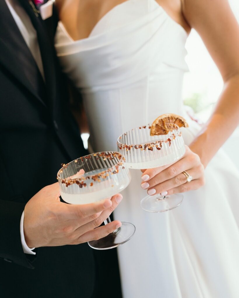 Close-up of bride and groom toasting signature cocktails garnished with citrus at a stylish Naples Florida wedding reception planned by expert wedding planners Naples FL and documented by photographer Priscila del Cristo.