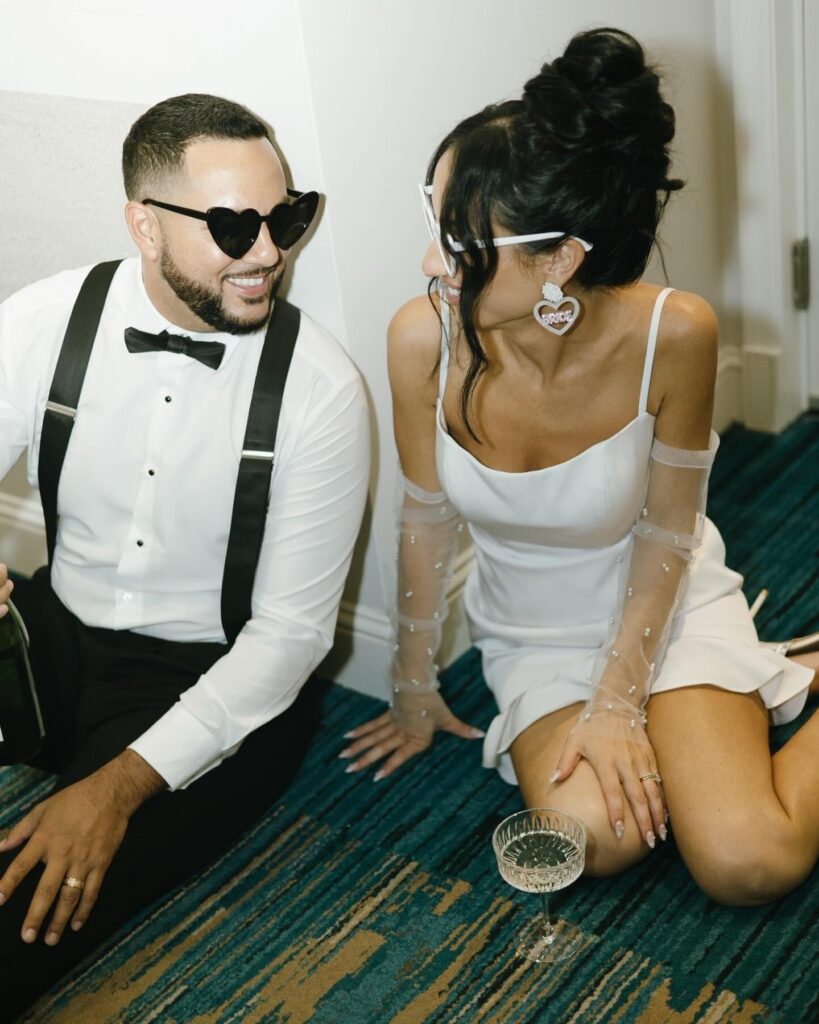 After-party portrait showcases the bride and groom relaxing and laughing together, fully embracing the joyful energy of their wedding celebration. Seated side by side, they exchange affectionate smiles while wearing matching heart-shaped sunglasses, adding a fun and modern twist to traditional wedding imagery. The bride’s sleek mini dress with sheer embellished sleeves and bold bridal accessories reflects a contemporary reception look, while the groom’s suspenders and bow tie maintain classic elegance. A cocktail glass nearby hints at the ongoing celebration, capturing a candid, stylish moment that perfectly reflects personality, connection, and post-ceremony bliss.