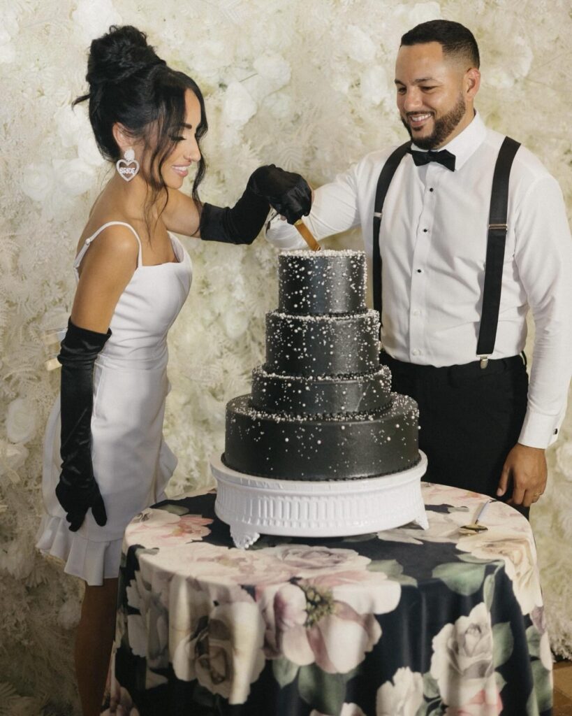 A stylish and fashion-forward cake-cutting moment captures the couple celebrating in front of a luxurious white floral backdrop. The bride wears a modern short white reception dress paired with elegant black opera gloves and statement heart-shaped earrings, creating a chic editorial look. The groom complements her style in a classic white dress shirt with black suspenders and a bow tie. Together, they smile as they slice into a dramatic multi-tier black wedding cake adorned with delicate pearl-like detailing, blending modern sophistication with playful personality. The scene feels intimate, celebratory, and effortlessly glamorous.