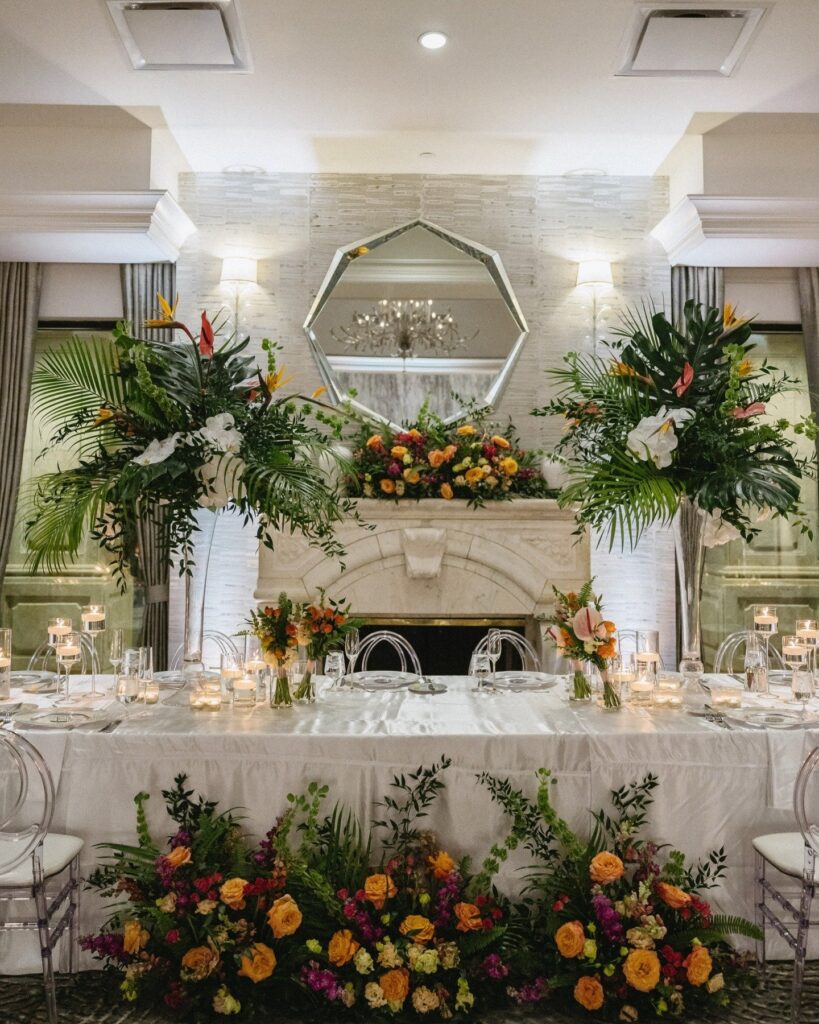 Elegant reception head table styled with lush tropical greenery, candles, and vibrant floral arrangements in front of a fireplace and mirror, highlighting luxury décor by wedding planners Naples Florida and a Naples Florida wedding planner team, photographed by Priscila del Cristo.