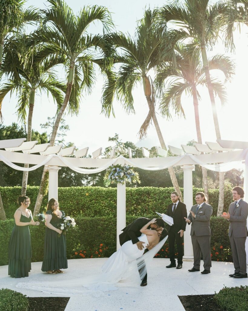 A joyful Naples Florida wedding ceremony unfolds beneath a white pergola draped with soft fabric and lush blue-and-white florals. The couple shares their first kiss in a romantic dip as guests and wedding party members celebrate around them. Framed by swaying palm trees and golden sunlight, this elegant outdoor wedding ceremony captures the beauty of a luxury destination wedding in Florida, highlighting thoughtful design, expert coordination, and timeless ceremony styling curated by a professional luxury wedding planner.