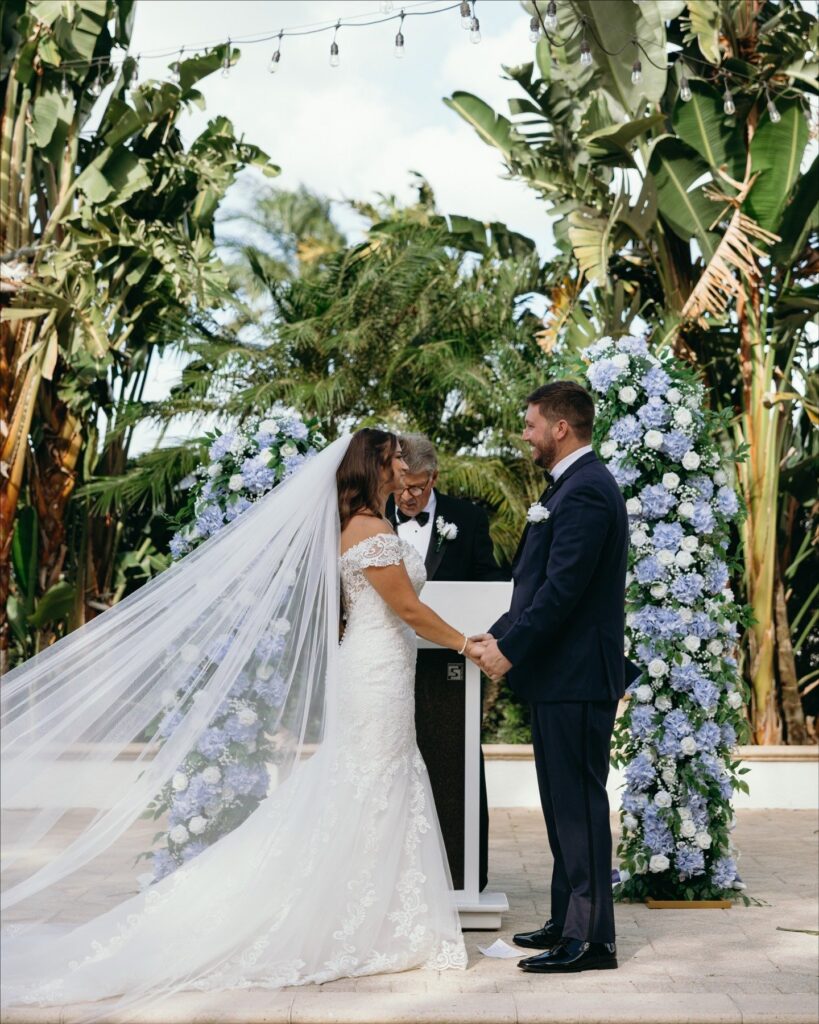 Bride and groom exchange vows beneath a floral ceremony arch with blue hydrangeas and lush greenery at a tropical outdoor venue, an elegant example of a Naples Florida wedding planner creating a romantic garden ceremony setting, photographed by Priscila del Cristo.