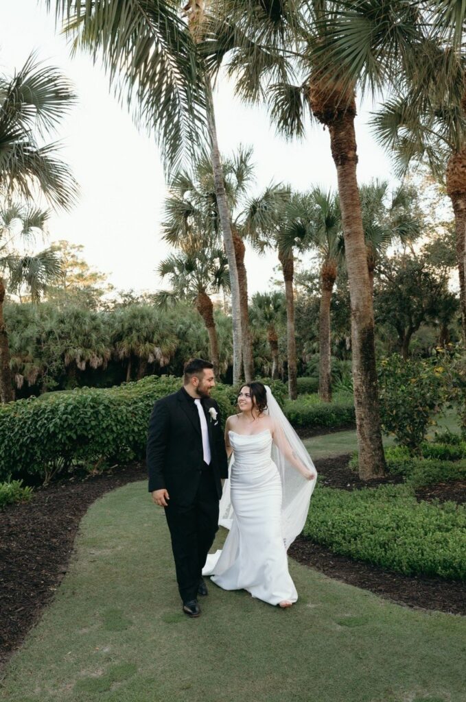 The newlyweds walk hand-in-hand through a tropical garden pathway, creating timeless bride and groom portraits in Naples FL. Surrounded by lush greenery and palm trees, the couple enjoys a quiet moment together during their luxury garden wedding celebration. Soft natural light enhances the romantic atmosphere, showcasing refined styling and elegant details that define elevated Florida wedding photography at a beautiful outdoor Naples wedding venue.