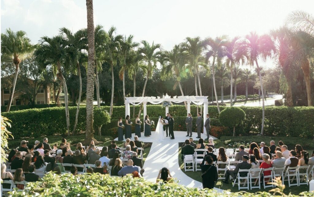 A sweeping perspective reveals the full outdoor ceremony design at a stunning Naples wedding venue, featuring a white aisle leading guests toward a floral-accented pergola altar. This panoramic view highlights intentional layout, guest experience, and cohesive event styling achieved through expert wedding planning in Naples Florida. The elegant ceremony setup provides beautiful destination wedding inspiration, blending natural surroundings with sophisticated luxury design.