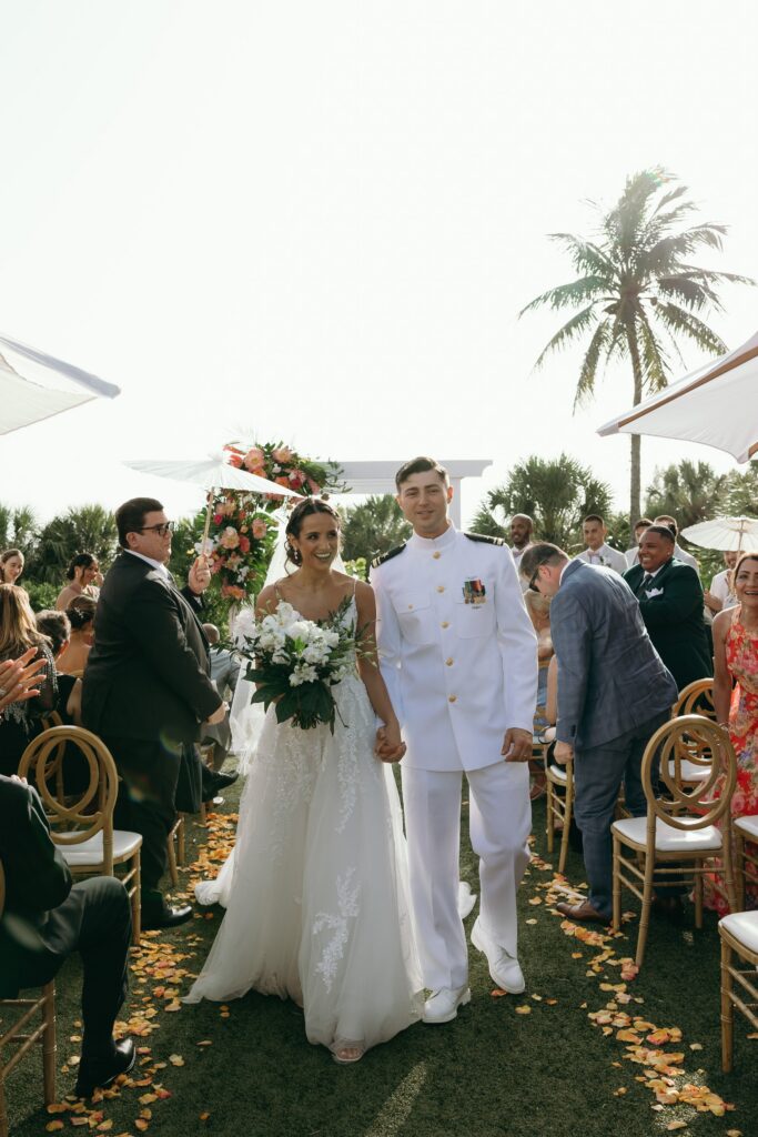 The bride and groom walk hand in hand down a grassy outdoor ceremony aisle in Naples, Florida, immediately after saying their vows. The groom wears a formal white military dress uniform adorned with medals, while the bride wears a flowing lace wedding gown with delicate straps and carries a bouquet of white flowers and greenery. Guests seated in gold chairs applaud on both sides, and flower petals scatter across the aisle beneath their feet. A floral arch and palm trees frame the ceremony space, showcasing the elegant outdoor setting often designed by wedding planners Naples Florida couples rely on for seamless waterfront celebrations.