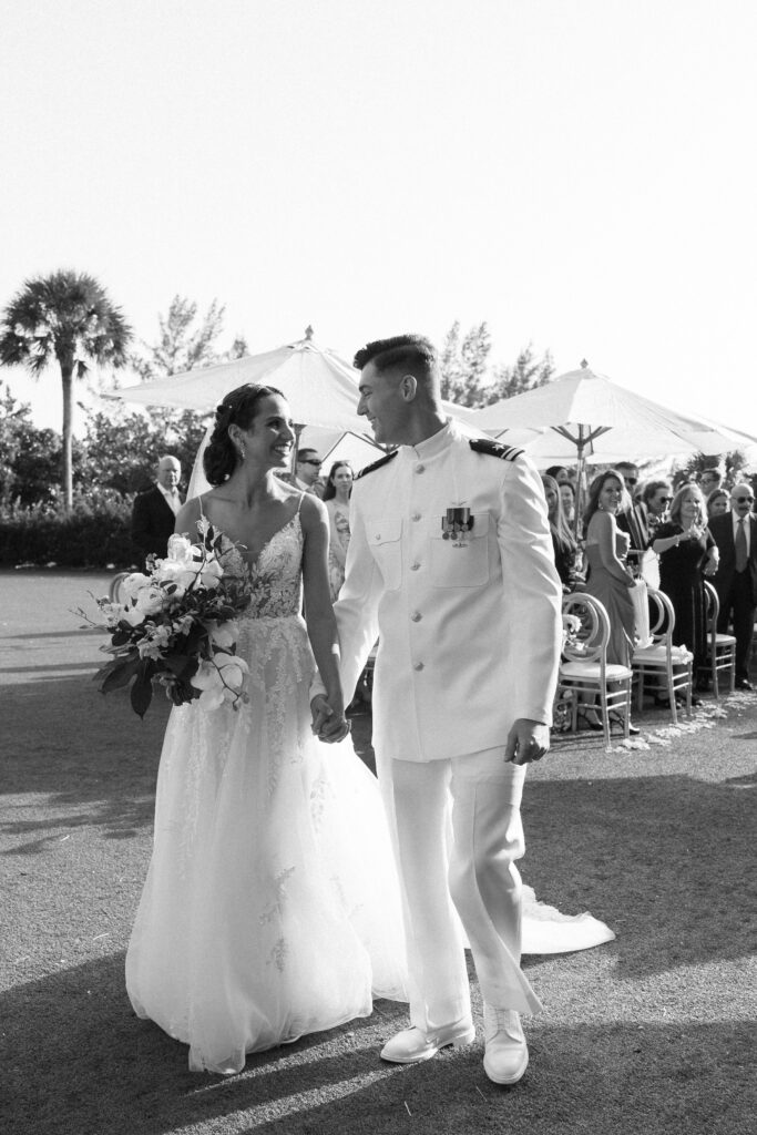 Black-and-white portrait of the bride and groom walking hand in hand across a sunlit lawn following their outdoor ceremony. The groom’s white military uniform contrasts sharply against the darker background of guests and palm trees, while the bride’s lace gown flows softly as she smiles up at him. Ceremony chairs and parasols are visible behind them, capturing a timeless moment that highlights the refined coordination and thoughtful styling a Naples Florida wedding planner brings to an elegant destination wedding.