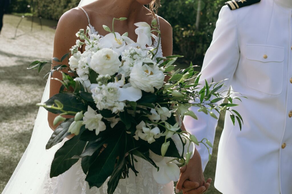 Close-up detail of a white bridal bouquet featuring orchids, ranunculus, and greenery as the bride and groom hold hands during a sunlit outdoor ceremony designed by wedding planner Naples FL professionals and photographed by Priscila del Cristo.