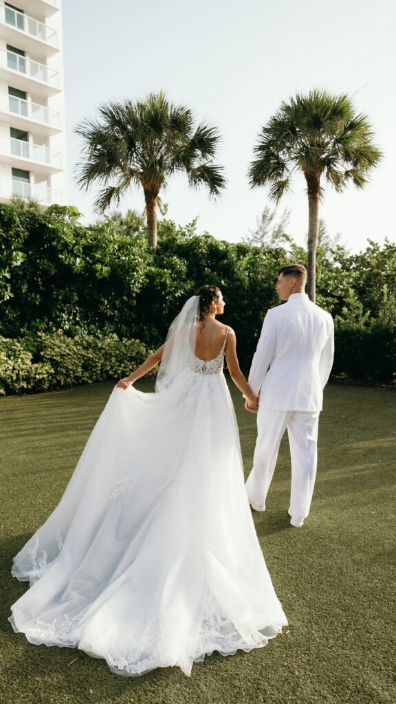 Back view of a bride and groom walking together beneath tall palm trees at a coastal venue, the bride lifting her long lace train as they stroll through a Naples FL outdoor ceremony space styled by experienced wedding planners Naples Florida and photographed by Priscila del Cristo.