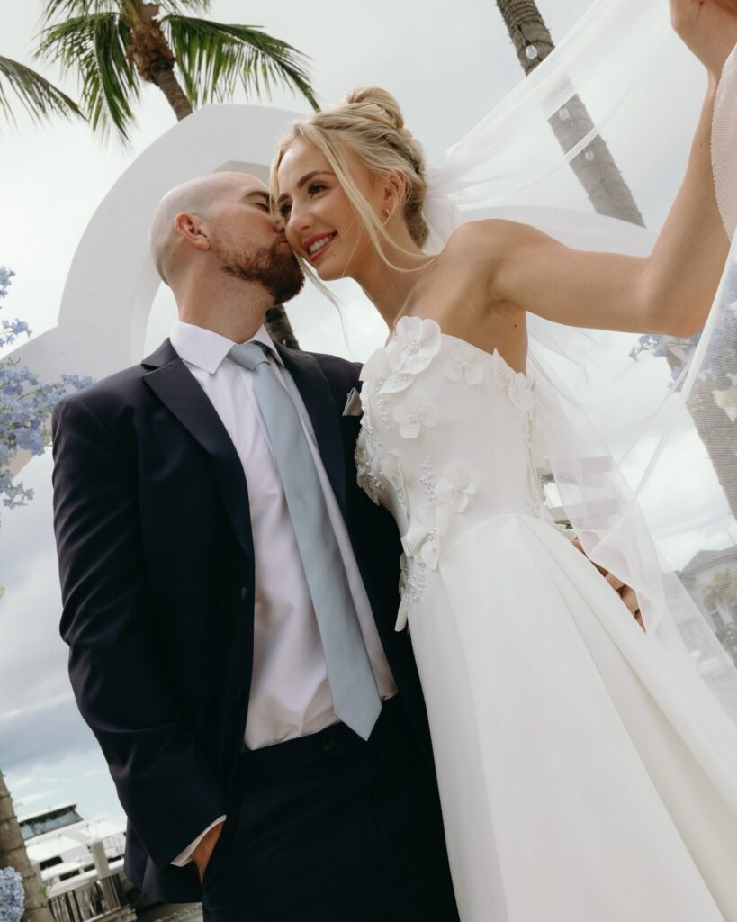 Set against the scenic Naples waterfront, this elegant ceremony features a grand floral arch adorned with soft blue and white blooms, framing the bride and groom as they celebrate their union. The sweeping bridal train and symmetrical ceremony design demonstrate the thoughtful coordination provided by an experienced Naples Florida wedding planner. From floral installations to guest sightlines and coastal styling, wedding planners Naples FL specialists create refined outdoor ceremonies that feel both luxurious and personal. This image reflects how wedding planner Naples FL professionals transform waterfront venues into unforgettable wedding experiences. Beautifully documented by Priscila del Cristo, the scene captures the sophistication and romance synonymous with weddings in Naples, Florida.