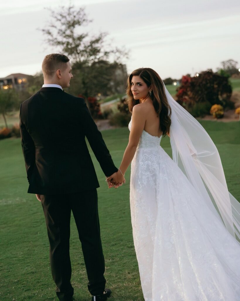 Bride and groom walk hand in hand across a green lawn at sunset during a romantic outdoor celebration designed by wedding planners in Naples, with the bride looking back over her shoulder as her long veil flows behind her, photographed by Priscila del Cristo for a Naples Florida wedding planner portfolio.
