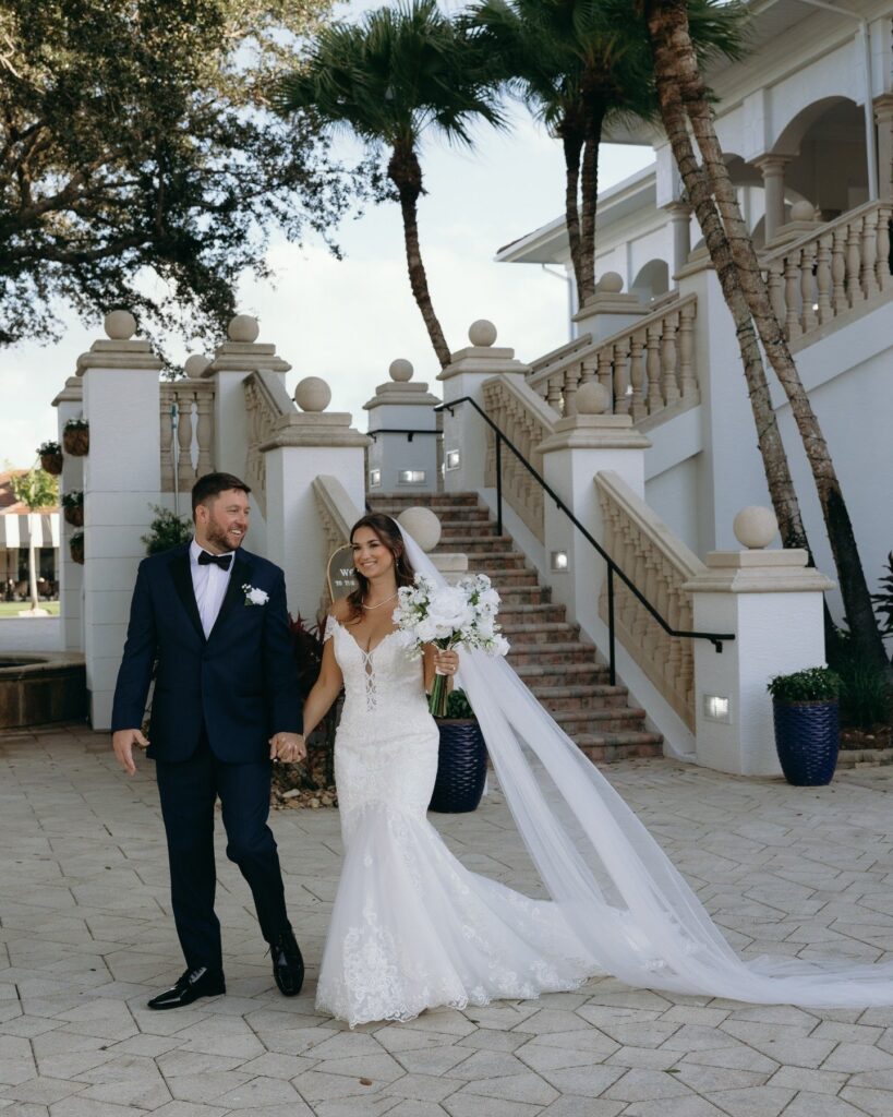 Bride and groom walk hand in hand outside a Naples Florida wedding venue, the bride wearing a fitted lace gown with a long flowing veil and carrying white roses as they smile beneath palm trees, beautifully coordinated by wedding planners in Naples.