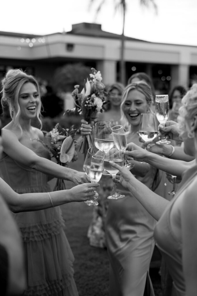 A group of bridesmaids gathers outdoors, laughing and clinking champagne glasses during a lively wedding celebration in Naples, Florida. The candid moment captures the excitement and connection shared among the bridal party as the festivities begin. Weddings like this highlight how wedding planners Naples FL seamlessly design relaxed yet elegant guest experiences, ensuring every celebration flows effortlessly from ceremony to reception. Photo by Priscila del Cristo.